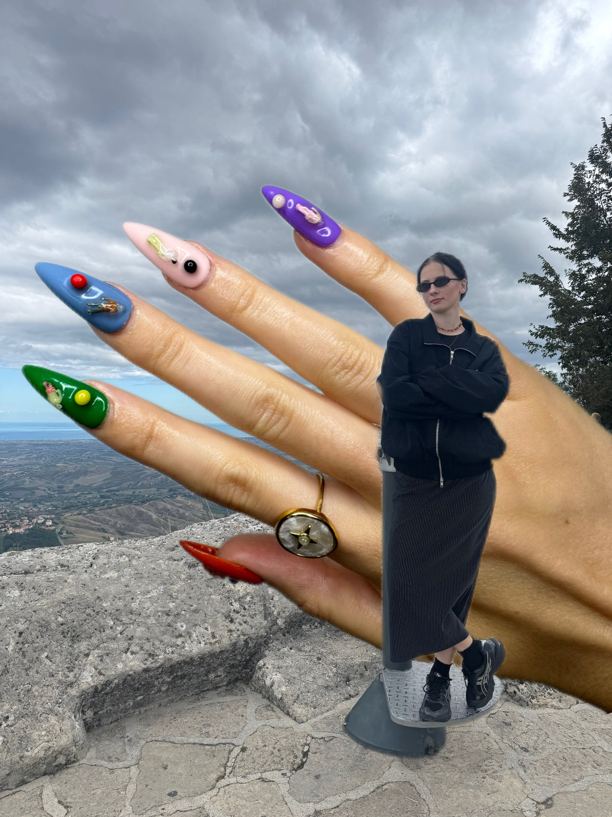 A person wearing a long black skirt, black jacket, and white sneakers stands on a stone cliff with a dramatic cloudy sky behind them. A giant hand with long decorated nails in blue, pink, purple, green, red, and gold is shown beside them.