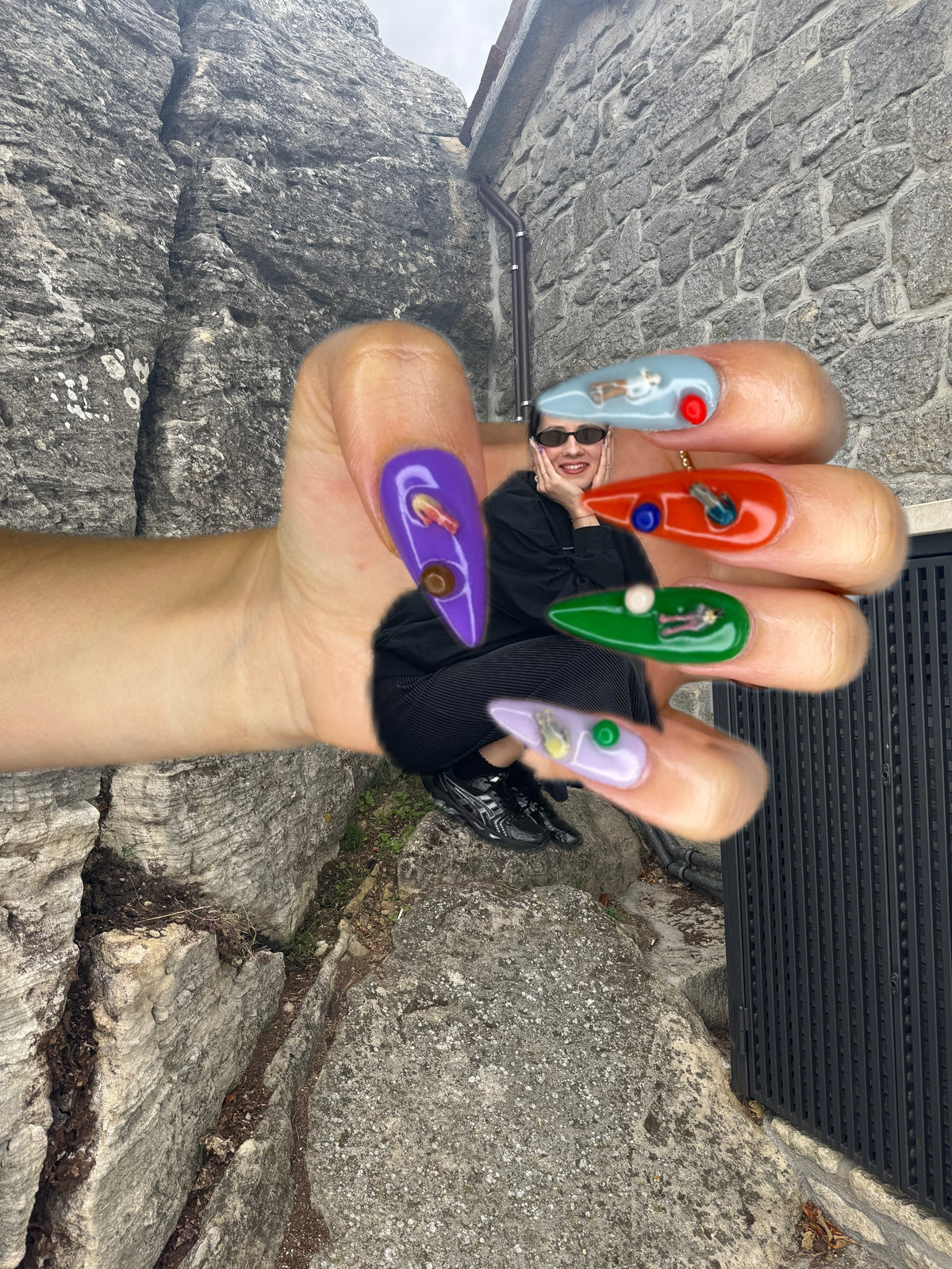 A hand with long, coffin-shaped nails decorated with colorful space-themed graphics (planets, stars, rockets) is shown in front of a stone wall background, with a person wearing sunglasses and a black outfit visible in the reflection.