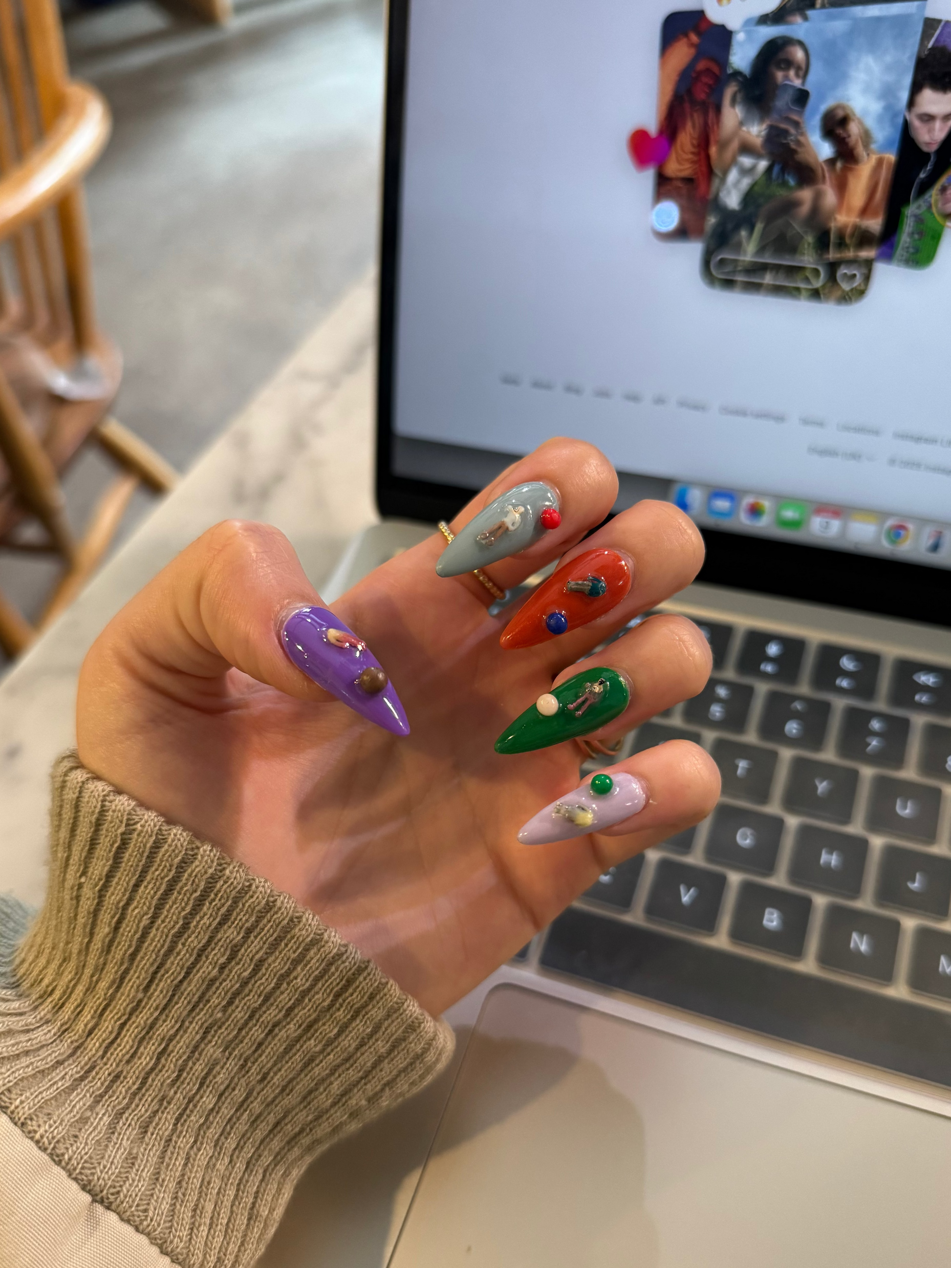 A hand with long, colorful stiletto-shaped press-on nails is shown in front of an open laptop. The nails are decorated in multiple vibrant colors including purple, orange, green, white, and silver, with small bead and glitter details.