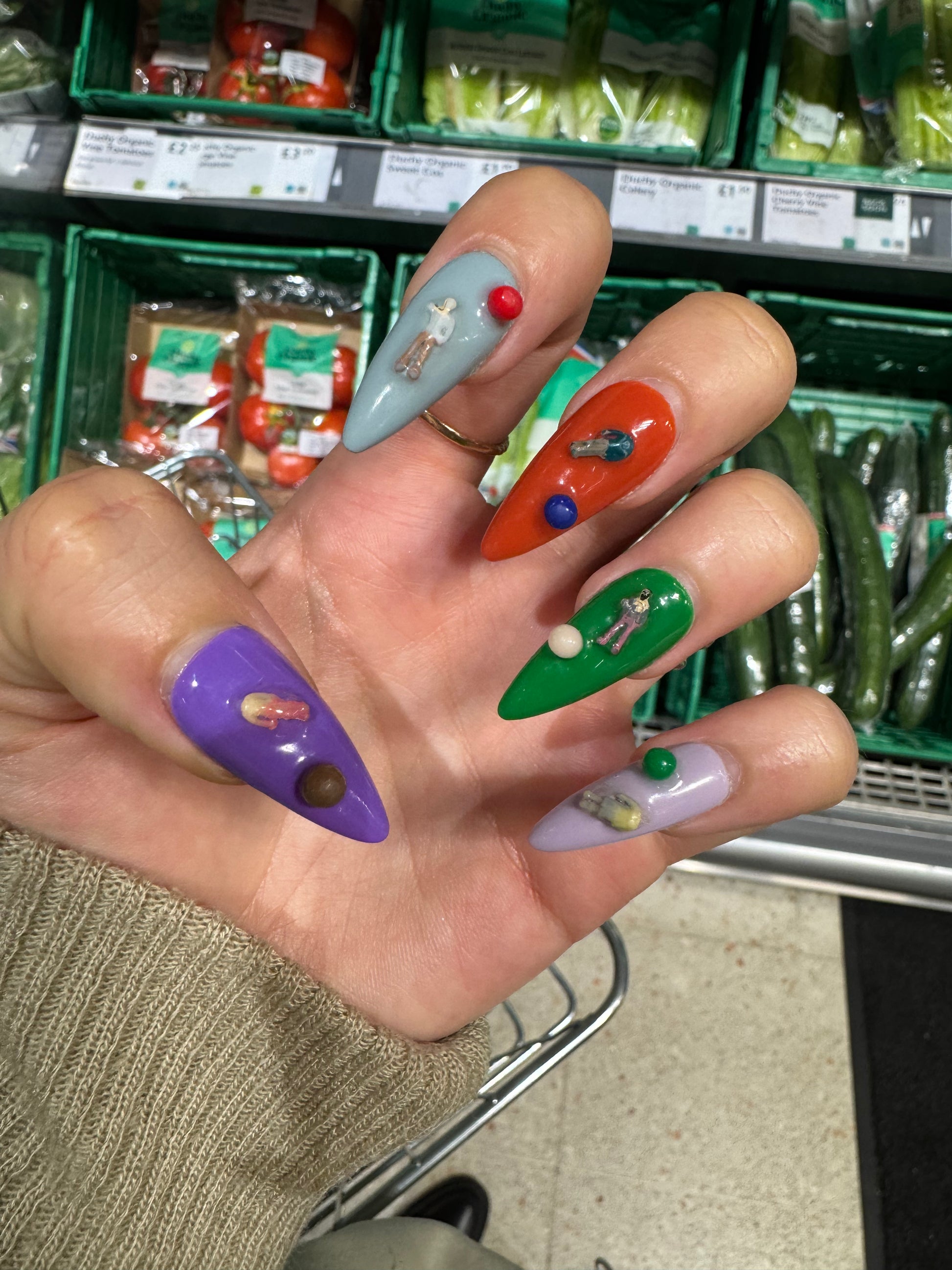A close-up of a hand with long, colorful stiletto-style nails decorated with small 3D figures and beads, photographed in a grocery store produce section.