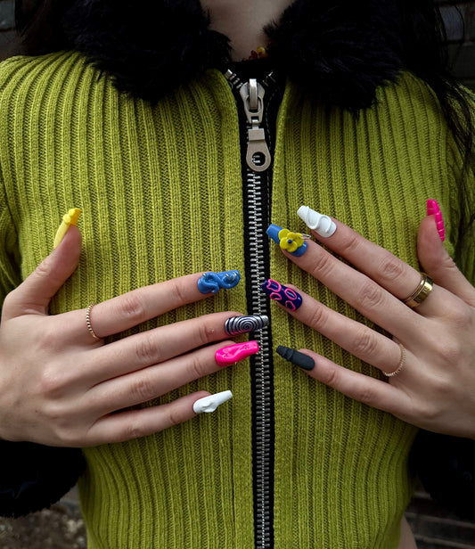 A close-up of a person wearing a bright olive green ribbed zip-up jacket with a black faux-fur collar, holding their hands in front of the zipper to display a variety of colorful, sculpted coffin-shaped press-on nails.