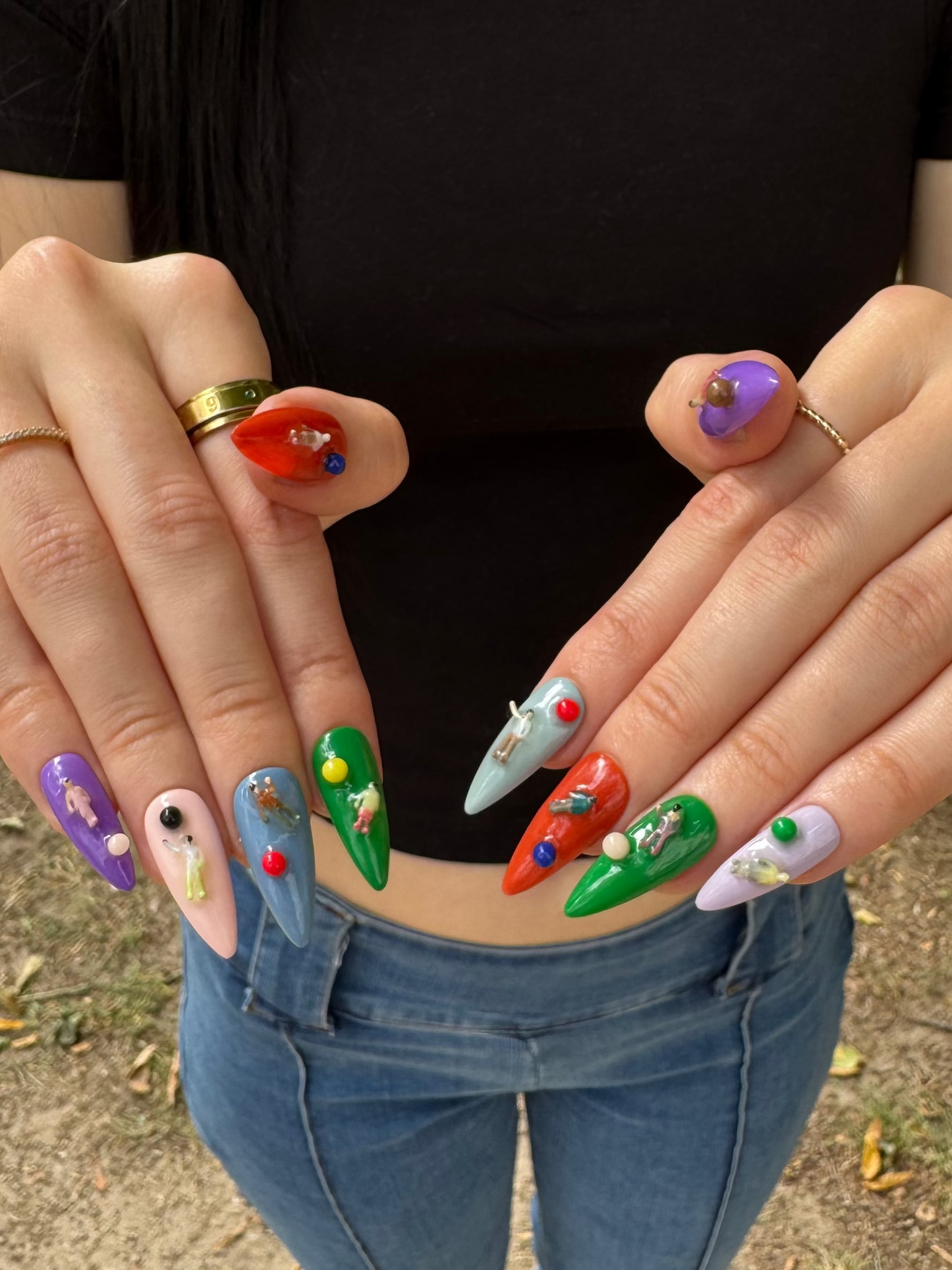 A person outdoors wearing a black t-shirt and blue jeans, showcasing a set of long, stiletto-shaped press-on nails with colorful, whimsical designs including green pumpkins, red fire trucks, purple flowers, and a light blue airplane.